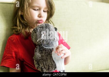 Kleines Mädchen hält Inhalationsmaske zu Hause. Lustige Kinder spielen mit Spielzeug und Vernebler. Baby mit Geräten zur Behandlung von Asthma oder Bronchitis. Konzept der Behandlung Stockfoto