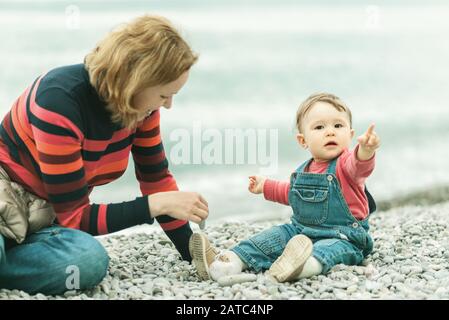 Süßes Baby und seine Mutter spielen mit Kieselsteinen. Die glückliche Familie ist im Frühling am Strand. Ein einjähriges Kind hebt sich die Hände. Verspieltes schönes Kleinkind in de