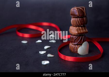 Stapel von Schokoladenbonbons und rotem Band auf schwarzem Hintergrund. Fokus auf Schokolade. Stockfoto