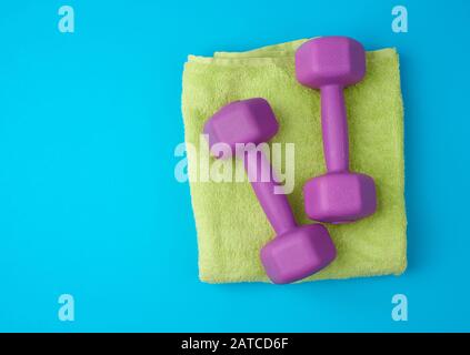 Ein Paar lilakische Kunststoff-Kurzhanteln liegen auf einem hellgrünen Frotteehtuch, blauer Hintergrund aus einem Teppich, Kulisse für Sport, Kopierraum Stockfoto