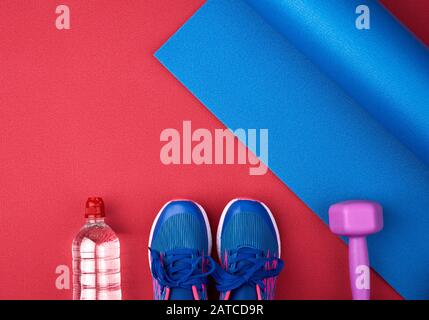 Zwei blaue Turnschuhe mit Schnürsenkeln, einer Flasche Wasser und einer blau verdrehten Neopren-Matte auf rotem Texturhintergrund, Draufsicht, Sportset Stockfoto