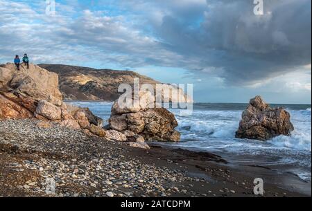 Zwei Touristen Frauen in Winterkleidung sitzen auf einem Felsen und genießen die stürmische Meereslandschaft. Felsen der Aphrodite in Zypern Stockfoto