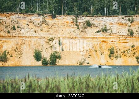 Hohe felsige sandige Steilufer mit Pinien bedeckt über einem klaren und blauen See. Schnelle Motorboot schneidet durch die Wasseroberfläche Stockfoto