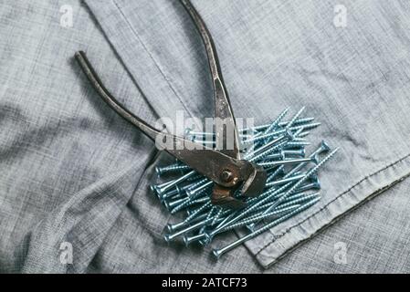 Eine Handvoll metallischer selbstschneiderschrauben und sehr gealterte und rostige Zangen liegen auf dem Hintergrund eines alten Denim-Stoffes. Makro mit selektivem Fokus aufgenommen mit Stockfoto