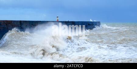 Eine Hafenmauer mit einem rauhen stürmischen Meer, das gegen die Mauer kragt und das Meer verwischt und in Bewegung versetzt, dahinter befindet sich ein rot-weißer Leuchtturm, wav Stockfoto