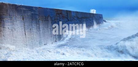 Eine Hafenmauer mit einem rauhen stürmischen Meer, das gegen die Wand kragt und das Meer verwischt und in Bewegung ist, stürzen Wellen über die Wand, dort i Stockfoto
