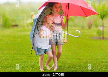 Zwei süße kleine Schwestern in einer Pfütze holding Schirme an einem regnerischen Sommertag stehend Stockfoto