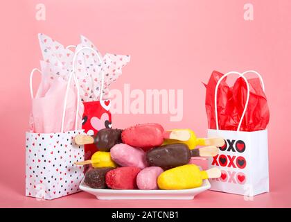 Bunte Süßigkeiten-bedeckte Kuchenpopskel auf einem weißen Teller, die abwechselnd Aromen und Richtung der Kuchenpops zeigen, auf einem rosafarbenen Hintergrund mit Valentinstag Geschenk Stockfoto