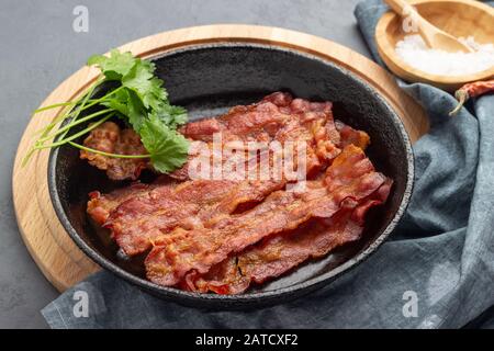 Knuspriger gebratener Speck in einer gusseisernen Pfanne auf grauem Grund. Stockfoto