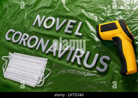 Wörter Roman Coronavirus mit Metallbuchstaben auf grün zerknittert gelegt Plastikfolie Hintergrund mit Gesichtsmasken und Thermometer Stockfoto