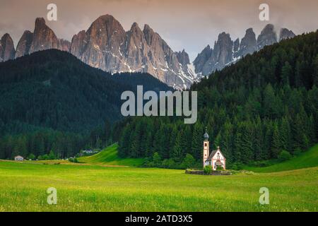 Beliebte Reiselage, süße alpine St. Johann Kirche im Tal des Val di Funes mit grünen Feldern und hohen Bergen bei Sonnenuntergang, Santa Maddalena touristisch Stockfoto