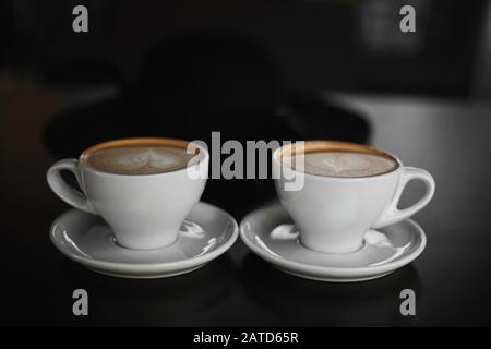 Zwei weiße Tassen Cappuccino-Kaffee mit herzförmigem Milchschaum. Seitenansicht auf zwei Tassen Latte Kaffee mit Herzfigur auf Milchschaum auf Schwarz Stockfoto