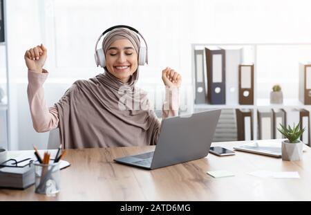 Fröhliche Muslimische Geschäftsfrau Hört Musik In Drahtlosen Kopfhörern, Sitzt Am Arbeitsplatz Stockfoto