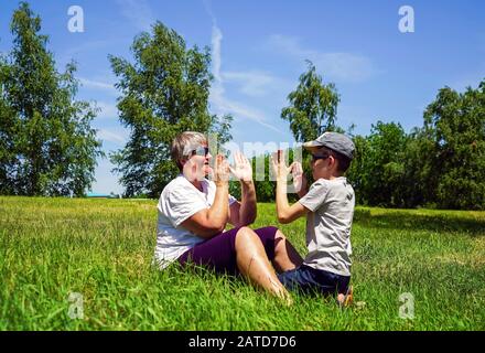 Ein Junge und eine Großmutter sitzen auf dem Rasen und spielen ein Spiel von Ladushki. Stockfoto