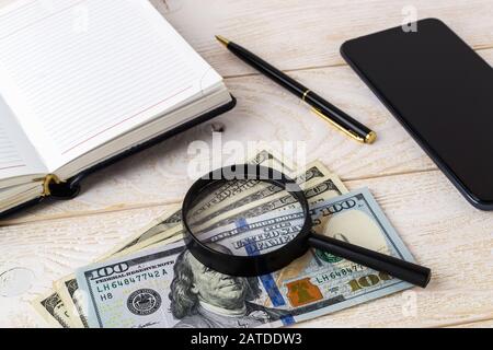 Lupe auf Dollarscheinen in der Nähe von Stift, Notizbuch und Smartphone auf einer weißen Holzoberfläche. Überprüfen Sie die Echtheit von Geld und Geschäftskonzept. Stockfoto