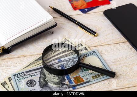 Lupe auf Dollarscheinen in der Nähe von Stift, Notizbuch, Kreditkarten und Smartphone auf einer weißen Holzoberfläche. Authentizität von Geld und Geschäftskonzept. Stockfoto