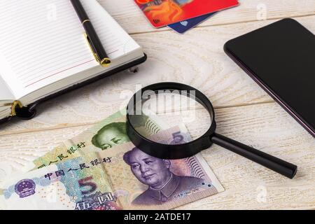 Lupe auf chinesischem Yuan in der Nähe von Stift, Notizbuch, Kreditkarten und Smartphone auf einer weißen Holzoberfläche. Authentizität von Geld und Geschäftskonzept. Stockfoto