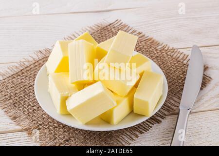 Rechteckige Stücke frische gelbe Butter auf einer weißen Untertasse und Tischmesser über einem weißen Holztisch. Molkereiprodukte. Sandwichbutter. Natürlicher Fettnährstoff. Stockfoto