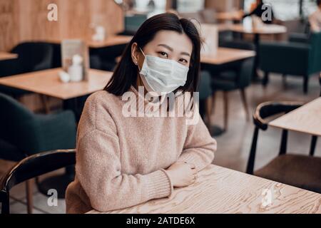 Asiatische Frau in einer Schutzmaske sitzt in einem Café. Schutz der Bevölkerung vor Viren durch Schutz der Atemwege. Das Konzept von coro Stockfoto