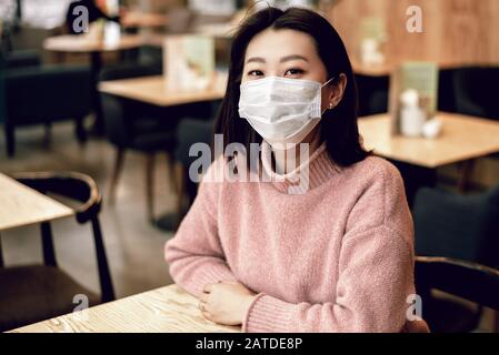 Asiatische Frau in einer Schutzmaske sitzt in einem Café. Schutz der Bevölkerung vor Viren durch Schutz der Atemwege. Das Konzept von coro Stockfoto