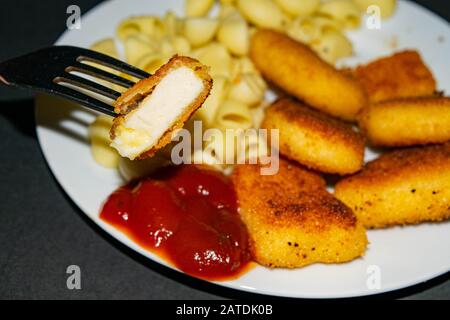 Ein Stück Nuggets auf einer Gabel und ein weißer Teller mit gebratenen Hühnerhuhn Nuggets, Pasta und Ketchup auf dunklem Hintergrund. Nahaufnahme Stockfoto