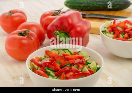 Vitaminsalat aus reifen Tomaten, frischen, grünen Gurken und roten Paprika in glasweißer Schüssel auf hellem Holztisch. Zutaten. Gemüse. Gesundheit Stockfoto