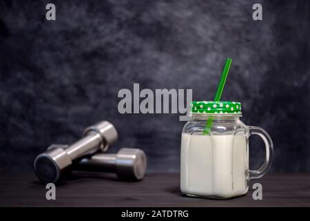 Weißer Cocktail in einem Becher mit Hanteln auf dunklem Hintergrund. Hintergrund. Konzept der Sporternährung und Proteindiät. Stockfoto