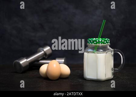 Eiweißcocktail in einem Becher und Hühnereiern auf dunklem Hintergrund mit Hanteln. Hintergrund. Konzept der Sporternährung und Proteindiät. Stockfoto