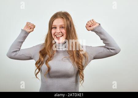 Eine junge fröhliche, rothaarige Frau im grauen Pullover ist glücklich und hat ihre Hände angehoben Stockfoto