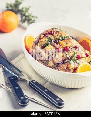 Gebratenes Schweinefleisch in weißem Gericht, weihnachtlicher gebackener Schinken mit Preiselbeeren, Tangerinen, Thymian, Rosmarin, Knoblauch auf leichter Tischoberfläche Stockfoto