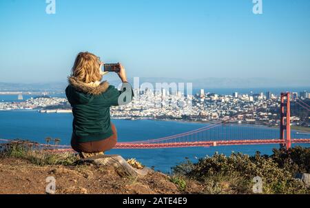 Junge blonde Frau in grüner Jacke, die mit ihrem Telefon der Golden Gate Bridge in San Francisco, Kalifornien, ein Foto macht. Tourist Foto von Golden Ga Stockfoto