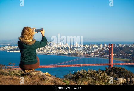 Junge blonde Frau in grüner Jacke, die mit ihrem Telefon der Golden Gate Bridge in San Francisco, Kalifornien, ein Foto macht. Tourist Foto von Golden Ga Stockfoto