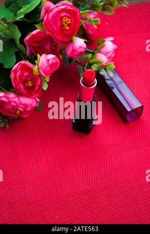 Roter Lippenstift mit Kappe, roten Blumen, rotem Hintergrund. Valentinstag. Geburtstag. Frauentag. Hen-Party. Festlicher Hintergrund. Kosmetik. Stockfoto