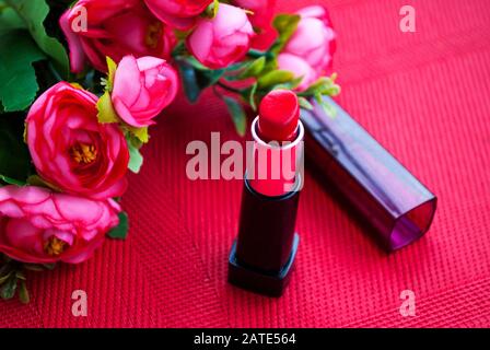 Roter Lippenstift mit Kappe, roten Blumen, rotem Hintergrund. Valentinstag. Geburtstag. Frauentag. Hen-Party. Festlicher Hintergrund. Kosmetik. Stockfoto