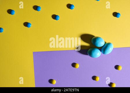 Von oben betrachtet schokolade Erdnusskugeln in bunter knuspriger Zuckerschale und drei blaue Eier isoliert auf gelbem und violett-geometrischem Hintergrund. Nahaufnahme von hübschen Süßigkeiten, die auf dem Tisch liegen, Schatten. Stockfoto