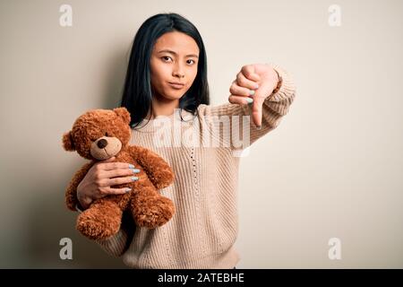 Junge, schöne chinesin, die Teddybär über isoliertem weißem Hintergrund hält, mit wütendem Gesicht, negatives Zeichen, das mit Daumen ablehnt Stockfoto