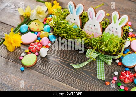 Selektive Fokussierung der mit osterginger glasierten Plätzchen in Form von Tieren, Eiern, drei Häschen in Schachtel mit gefälschtem Gras, Narzissen isoliert auf Holzhintergrund. Erdnussschokolade in der Zuckerschale. Stockfoto