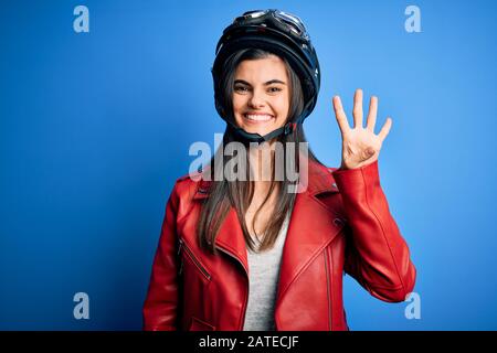 Junge schöne brünette Motorradfrau mit Motorradhelm und Jacke, die mit den Fingern Nummer vier zeigt und zeigt, während sie lächelnd konfi Stockfoto