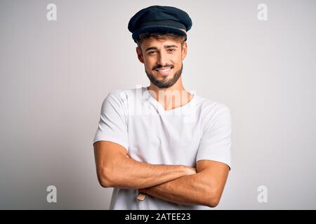 Junger Fahrer mit Bartehut, der über isoliertem weißem Hintergrund steht, glückliches Gesicht, das mit gekreuzten Armen lächelt, die auf die Kamera blicken. Positiv p Stockfoto