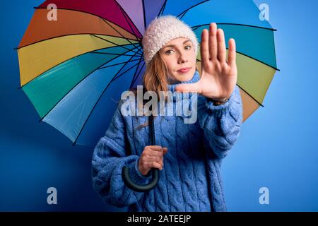 Junge, schöne blonde Frau, die einen farbenfrohen Regenschirm über isoliertem blauem Hintergrund hält, mit offenem Handzeichen und einem ernsthaften und selbstbewussten Ausdruck Stockfoto