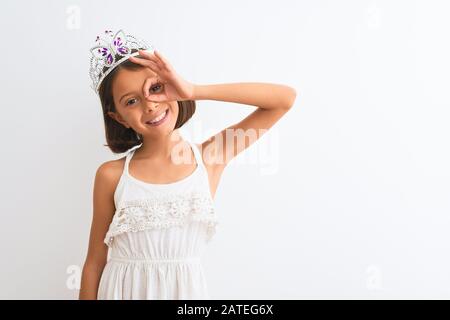 Schönes Kindermädchen, das die Fürstenkrone trägt und über einem isolierten weißen Hintergrund steht. Es geht mit der Hand, die lächelt, mit dem Auge, das durch die Finger schaut, gut Stockfoto