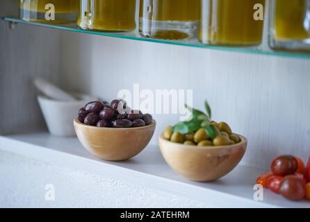 Stimmungsvolle Nahaufnahme von grünen und schwarzen Oliven in Holzschüsseln neben verschwommenem weißen Mörser und Tomaten, Glasflaschen mit Ölgehalt können Stockfoto