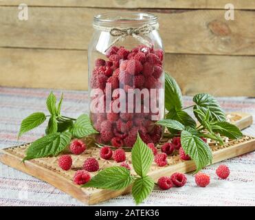 Himbeere in einem Glaskerl mit frischen Blättern und Beeren, die auf Holzschneidebrett auf rustikalem Hintergrund verstreut sind, Nahaufnahme, Kopierraum, gesund Stockfoto