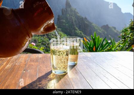 Man gießt Weißwein aus Ton Kanne in Glas auf der Terrasse mit Blick auf die grüne Landschaft der kleinen Bergdorf Masca auf Teneriffa, Spanien im sonnigen d Stockfoto