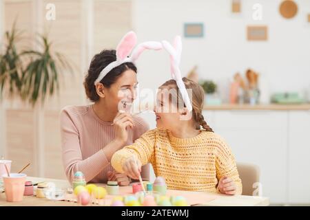 Porträt der liebenden Mutter mit kleinen Mädchen Spaß beim Malen von Ostereiern in gemütlicher Kücheneinrichtung, beide tragen Hunnenohren, Kopierraum Stockfoto