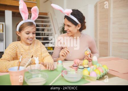 Porträt der glücklichen jungen Mutter und Tochter, die Ostereier im gemütlichen Kücheninneren malen, beide mit Hunnenohren, Kopierraum tragen Stockfoto