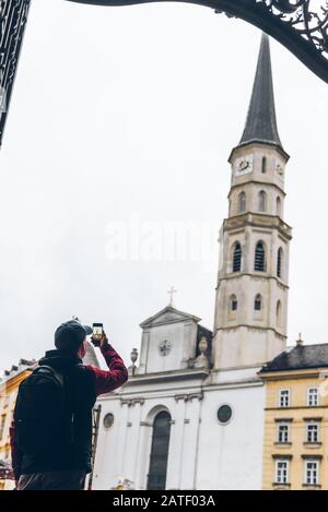 Mann Tourist im Regenmantel mit Rucksack und Blick auf die Kirche von Saint michaels, die am Telefon fotografiert Stockfoto