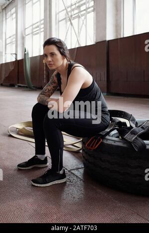 Junge, tätowierte kaukasische Frau, die in der Sporthalle auf dem Reifen sitzt Stockfoto