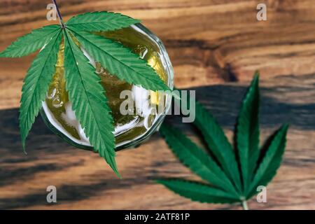 Eiskaltes Marihuana Cannabis infundiertes THC-Getränk in einem kristallklaren Glas mit Unkrautblättern auf dunklem Holzhintergrund. CBD-Medikamentengetränk Stockfoto