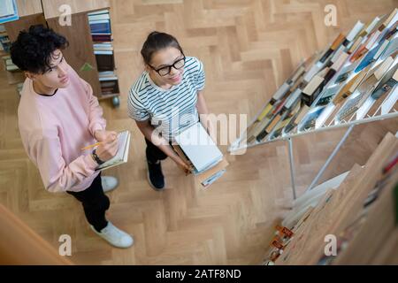 Zwei clevere Teenager-Studenten in Casualwear, die einen der Bücherregale betrachten Stockfoto
